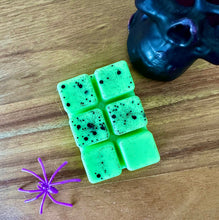 Load image into Gallery viewer, A green wax melt bar with black speckles, styled on a wooden surface next to a purple plastic spider and a black skull prop.